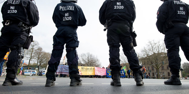 Polizisten in Montur stehen auf einer Straße. Am Horizont sind Demo-Banner zu sehen.