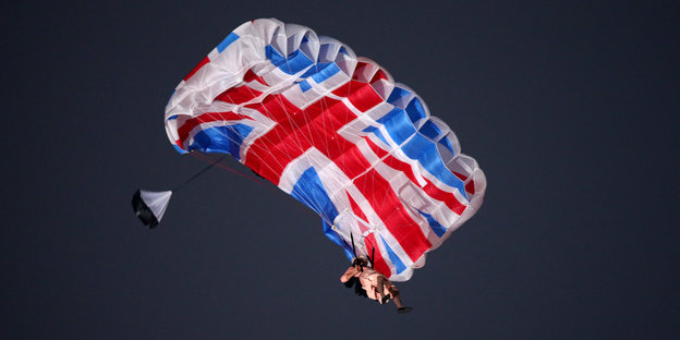 Ein als die Queen verkleideter Schauspieler hängt an einem Fallschirm mit britischem Flaggenmotiv in der Luft