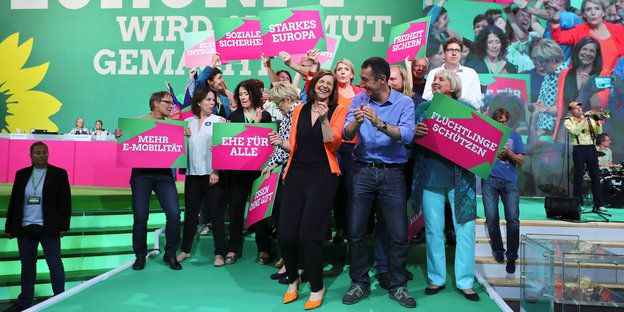 Viele Menschen stehen auf einer Bühne und halten Schilder mit den Zielen der Grünen in die Höhe. Sie lachen.