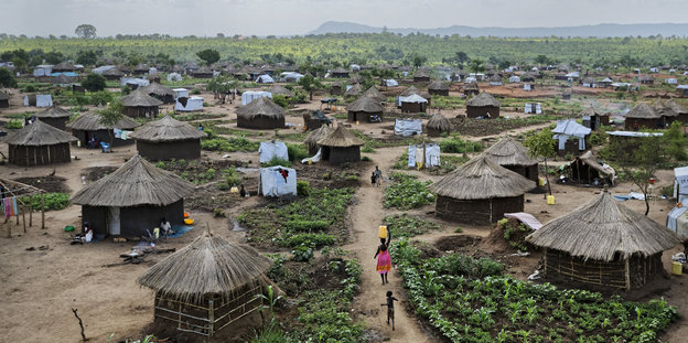 Häuser mit Strohdach stehen in einer idyllischen Landschaft. Dazwischen provisorische Zelthütten der UNHCR