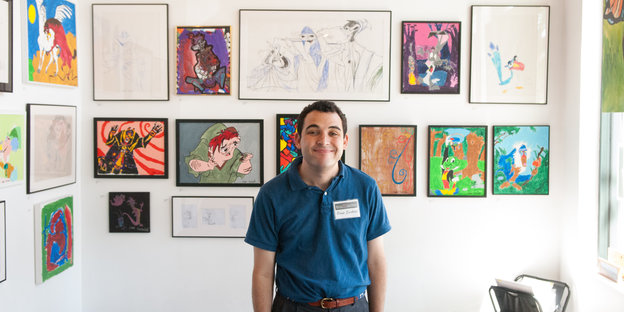 Ein junger Mann steht vor einer Wand voller Disney-Bilder und grinst
