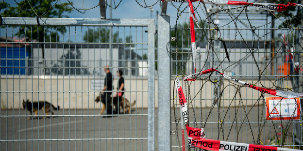 Polizist_innen mit Hunden hinter einem Gitter, das von Stacheldraht gesaäumt ist