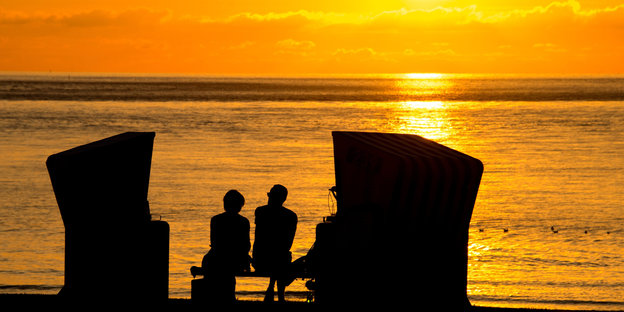 Zwei Menschen sitzen neben zwei Strandkörben am Meer, während die Sonne untergeht