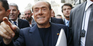 Berlusconi grinst dreist, zeigt mit dem Zeigefinger nach vorne und schreitet mit Paoieren unter dem Arm zwischen anderen Männern hindurch