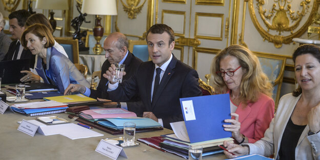 Emmanuel Macron sitzt zwischen seinen Kabinettsmitgliedern und hebt sein Wasserglas