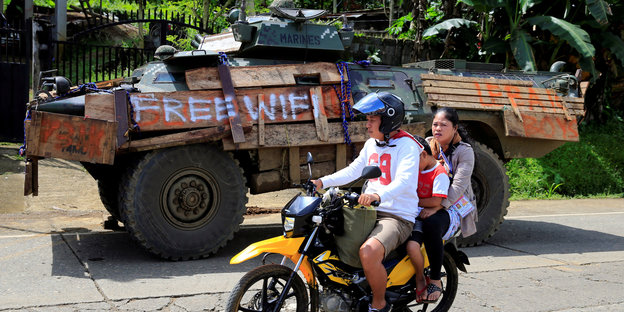 Panzer auf einer Straße, daneben fährt ein Moped mit einem Mann am Lenker und einer Frau auf dem Rücksitz