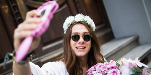 Ein junger Mann mit Sonnenbrille, langen Haaren und Blumenkranz filmt sich selbst mit einem pinken Handy