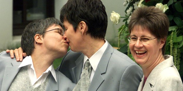 Zwei Frauen küssen sich, eine dritte steht daneben und lächelt