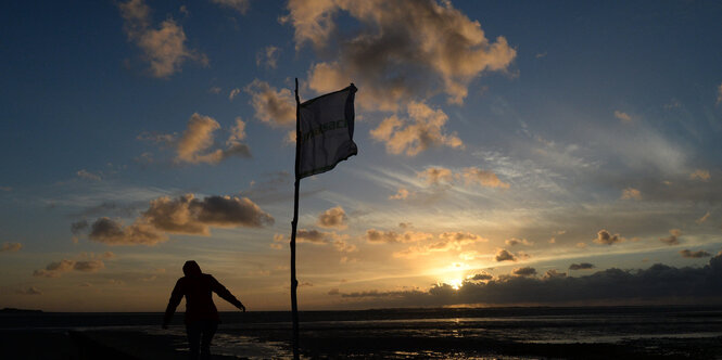 Eine Fahne weht im Wind, dahintertaucht die Sonne ins Meer, ein Mann schreitet durch die Dämmerung