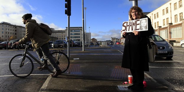 Eine Frau hält ein Schild in die Höhe, auf dem „Shame“ (Schande) steht, im Hintergrund ein Fahrradfahrer