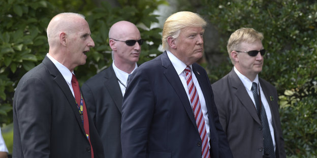 Donald Trump schreitet umringt von drei Leibwächtern durchs Bild