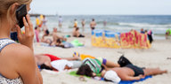 Eine Frau steht mit dem Rücken zum Betrachter am Strand und telefoniert, sie blickt auf schlafende Menschen in der Sonne