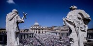 Zwei Statuen säumen den Blick auf den von Menschen gefüllten Petersplatz mit dem Petersdom im Hintergrund vor strahlend blauem Himmel