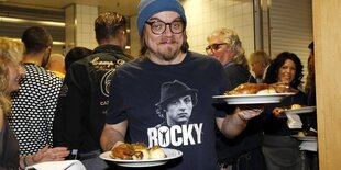 Mann mit Wollmütze, dicker Brille und "Rocky"-T-Shirt schneidet Grimasse und balanciert zwei Teller mit Essen