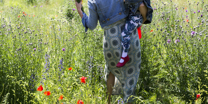 Eine Frau läuft mit ihrer Tochter auf dem Arm über eine Blumenwiese, fotografiert von hinten