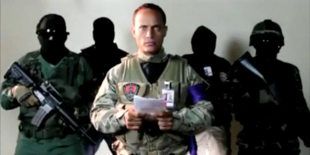 Ein Mann in Militärkleidung liest eine Botschaft vor, im Hintergrund maskierte Bewaffnete
