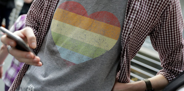 Ein Mann mit einem regenbogenfarbenen Herz auf dem T-Shirt tippt mit einer Hand auf seinem Handy