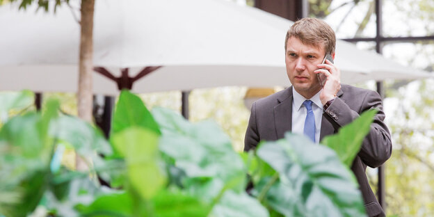 Andre Poggenburg steht mit Handy am Ohr hinter einer grünen Pflanze