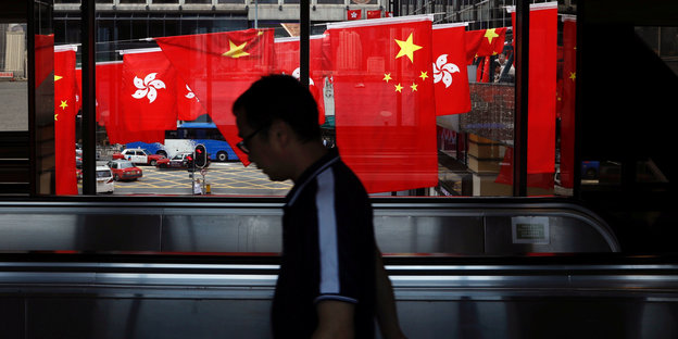 Ein Mann geht mit gesenktem Kopf über eine Rolltreppe, im Hintergrund hängen überdimensionale chinesische Flaggen
