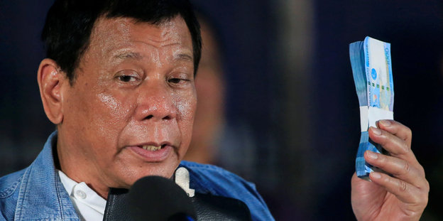 Rodrigo Duterte steht hinter einem Mikrofon und hält ein Bündel Geldscheine in die Höhe