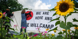 Menschen hängen ein Transparent auf, auf dem steht „We are here and we will camp“, davor eine große Sonnenblume