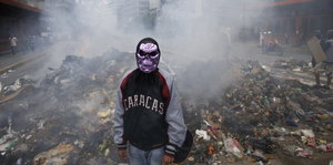 Ein Maskierter Demonstrant steht vor einem brennendem Müllhaufen