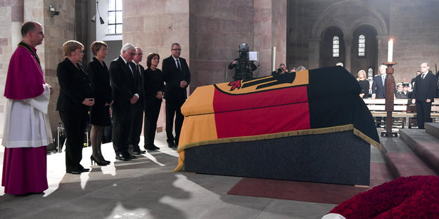 Ein mit deutscher Flagge umhüllter Sarg, davor stehen einige Menschen