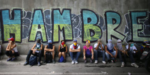 Menschen sitzen vor einer Wand mit der Aufschrift "HAMBRE"