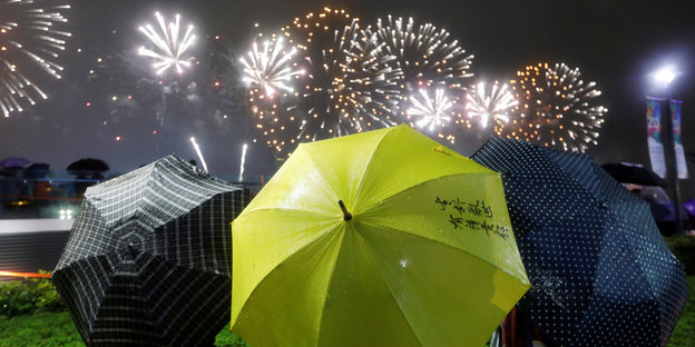 Drei Personen mit Regenschirmen sind von hintern zu sehen, im Hintergrund ein Feuerwerk