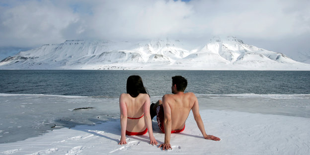 Zwei Menschen in Badekleidung sitzen im Eis und schauen auf einen Fjord