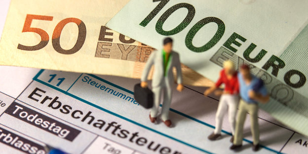 Man sieht ein Formular zur Erbschaftssteuererklärung, einen 50- und einen 100-Euroschein und zwei Modellfiguren