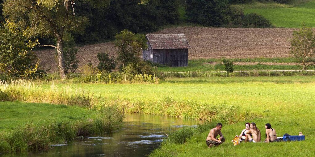 Menschen sitzen neben einem Bach in einer grünen Landschaft