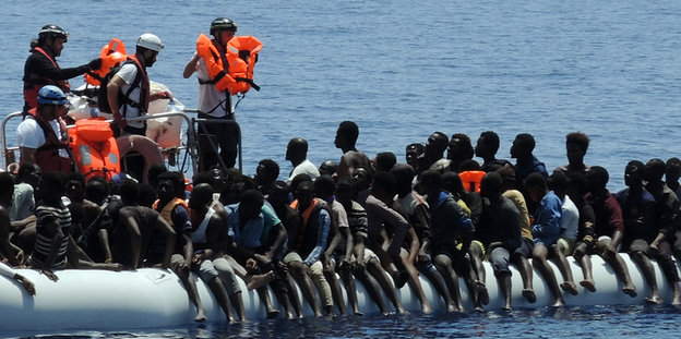 Migranten sitzen in einem überfüllten Schlauchboot auf dem Mittelmeer. Helfer von Hilfsorganisationen nähern sich ihnen und verteilen Schwimmwesten