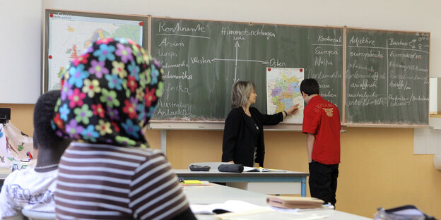 Ein Schüler und eine Lehrerin stehen an der Tafel, ein Mädchen mit Kopftuch schaut ihnen zu