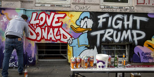 Ein Mann malt an einer Hauswand einj Graffito mit der Aufschrift "Love Donald, hate Trump" und einem Donald-Duck-Motiv