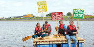 Vier Menschen auf einem Fluss. Sie halten Schilder in die Höhe, auf denen "soziale Gerechtigkeit" und "Demokratie" steht