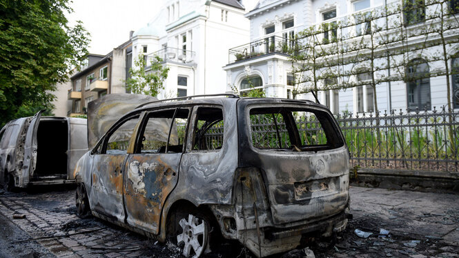 Ein ausgebranntes AUto in einer schicken Wohnstraße vor einem weißen Haus