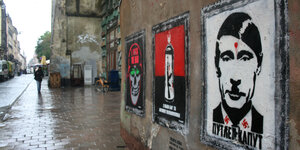 Plakate an einer Hauswand. Eines zeigt Putin mit einem Hitlerbärtchen und einer Kopfschusswunde