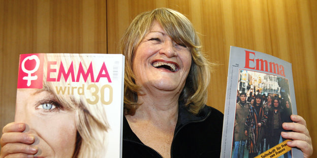 EIne Frau lacht und hält zwei Ausgaben der Zeitschrift Emma