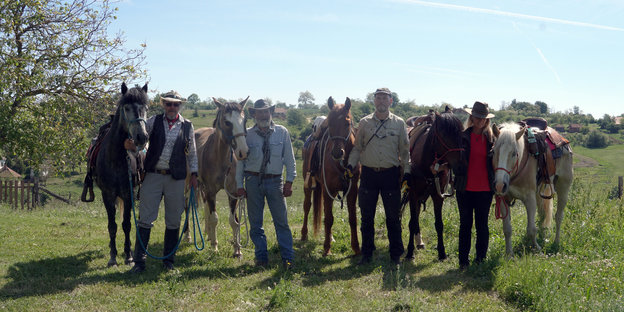 Fünf Pferde und vier Reiter posieren nebeneinanderstehend auf einer Wiese