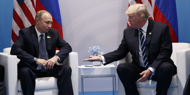 Zwei Männer sitzen im Sessel, es sind Putin und Trump, Trump streckt seine Hand zum Handschlag aus, Putin zögert sie zu nehmen