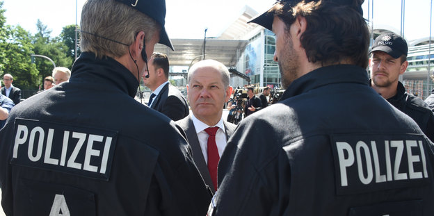 Olaf Scholz steht vor zwei Polizisten