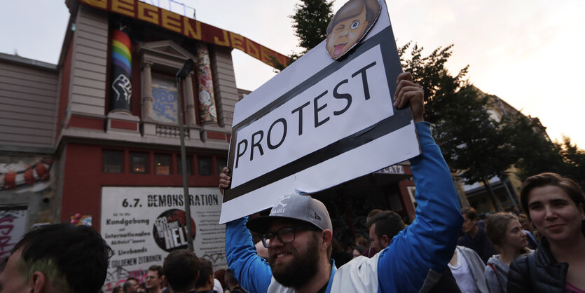 Ein Demozug vor der Roten Flora. Ein Mann hält ein Schild mit dem Schriftzug „Protest“ hoch