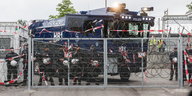 ein Wasserwerfer vor dem Polizisten stehen hinter einem Zaun mit Nato-Draht