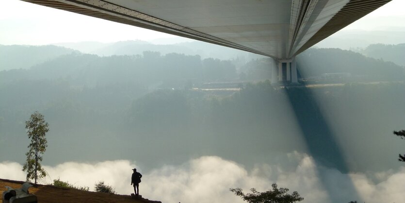 Unter einer Brücke über einen Fluß, am Ufer steht ein Mann. Das gegenüberliegende Ufer ist hügelig und bewaldet