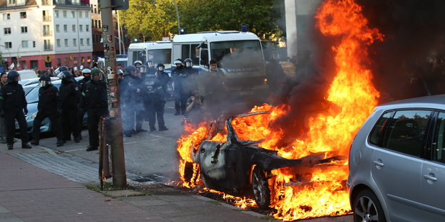 Ein brennendes Auto und Polizisten im Hintergrund