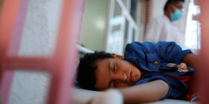 Ein Junge schläft auf einem Krankenbett. Hinter seinem Bett läuft ein Arzt entlang