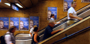 Personen auf einer Rolltreppe, im Hintergrund Poster mit dem Gesicht von George Soros