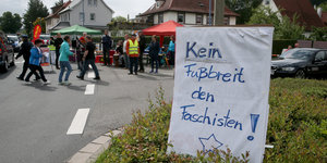 Auf einem Plakat steht „Kein Fußbreit den Faschisten“, dahinter Menschen und Sonnenschirme