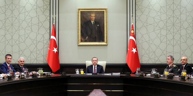 Erdogan und andere Männer sitzen unter türkischen Fahen und einem Porträt